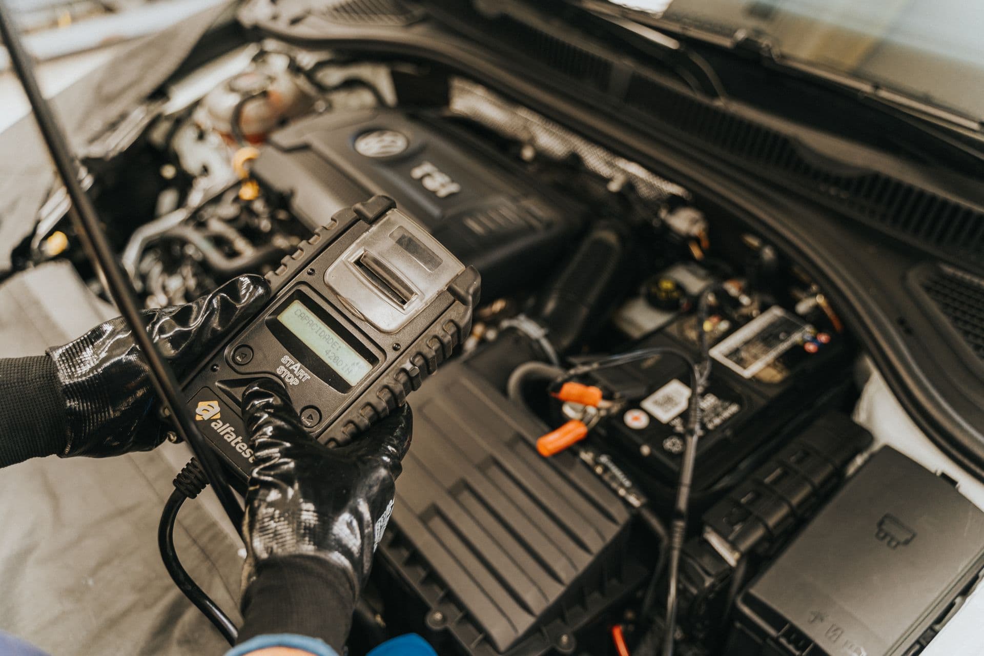 A technician holding a battery tester against a car battery in an open engine bay to read its condition