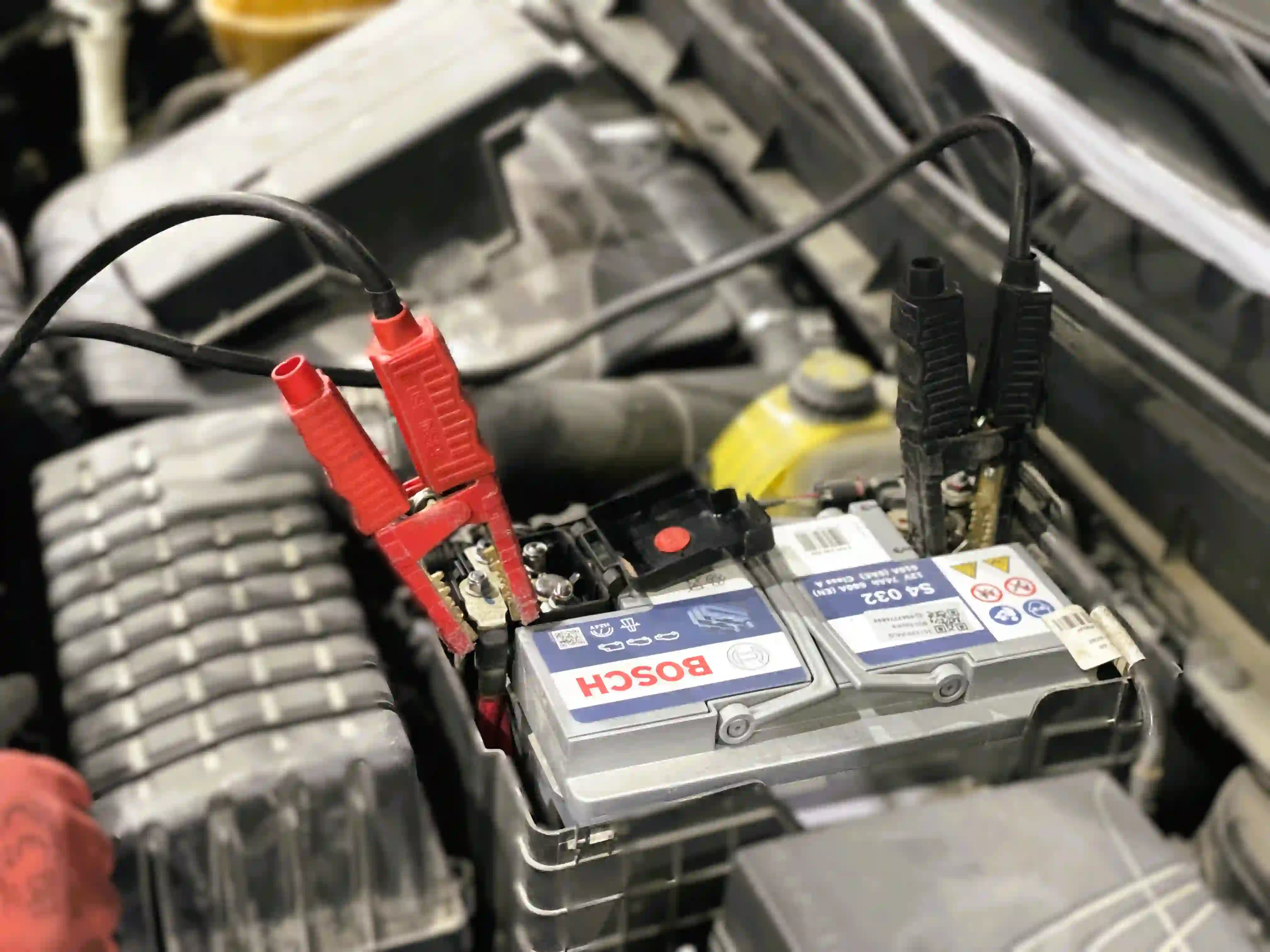 Red and black jumper clamps attached to the terminals of a Bosch S4 032 car battery, with its 12V 74Ah 680A rating printed on the label