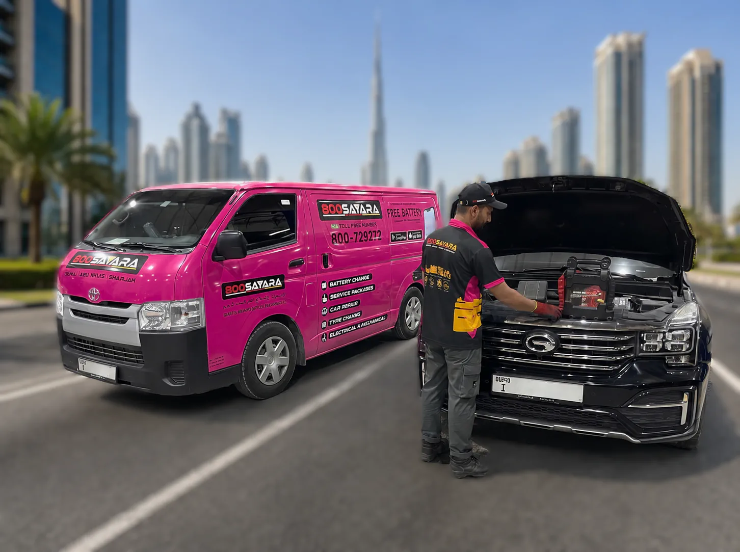A mobile battery service van parked beside a black SUV with its bonnet raised while a technician connects a portable jump starter, on a Dubai road with the Burj Khalifa and the city skyline behind