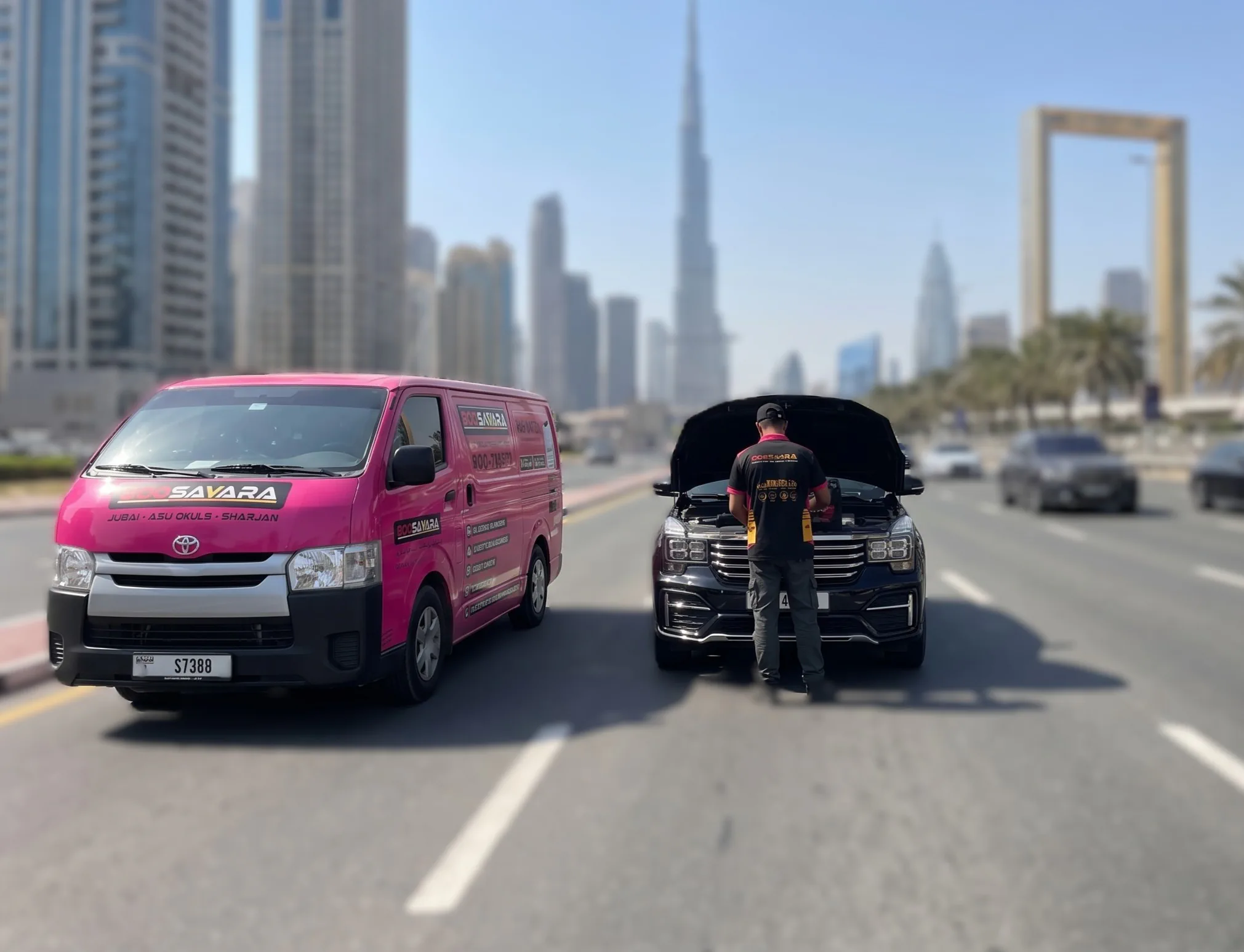 A mobile battery service van and a technician working under the raised bonnet of a stopped car on a Dubai road, with the Burj Khalifa and the Dubai Frame in the background